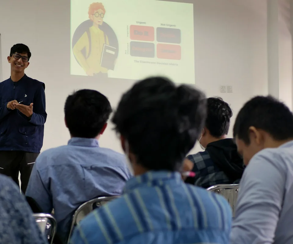 Pembicara berdiri di depan layar presentasi, peserta duduk mengikuti sesi